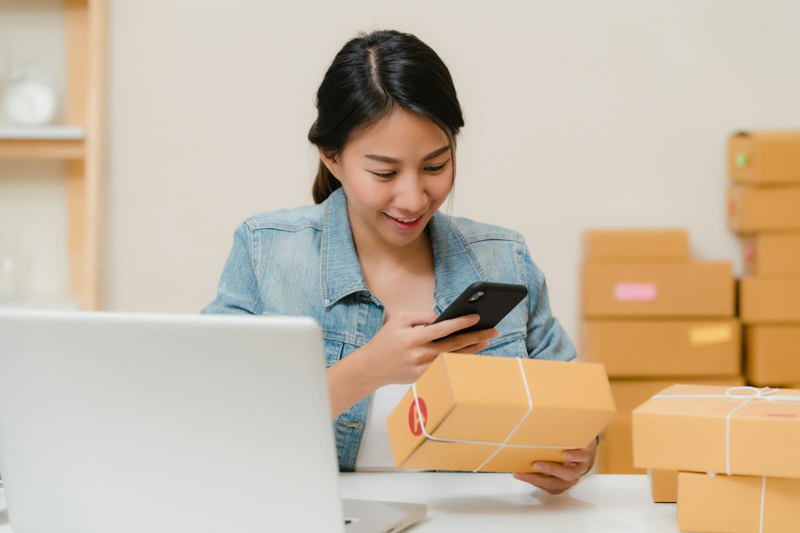 A small business owner smiling at her phone while checking an order, packed boxes stacked behind her